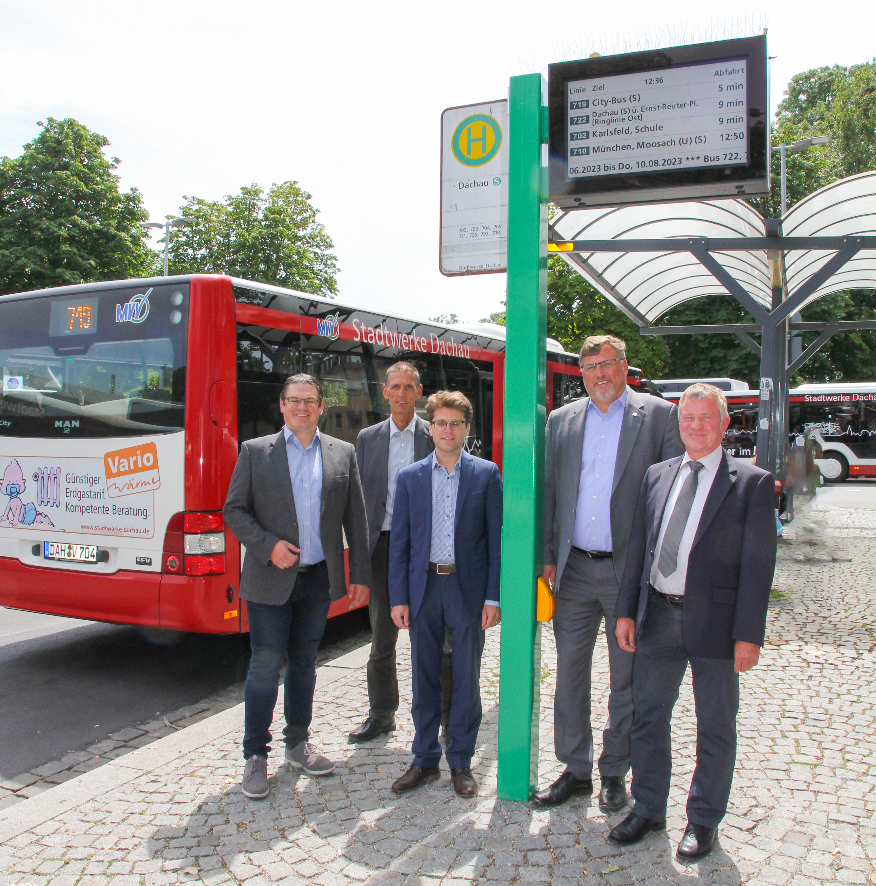 Menschengruppe steht unter dem neuen digitale Fahrgastanzeiger, im Hintergrund steht ein Bus