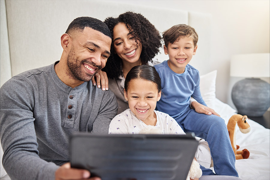 Eine glückliche Familie sitzt zusammen auf einem Bett und schaut auf ein Tablet. Die Eltern und zwei Kinder lächeln und genießen die gemeinsame Zeit.